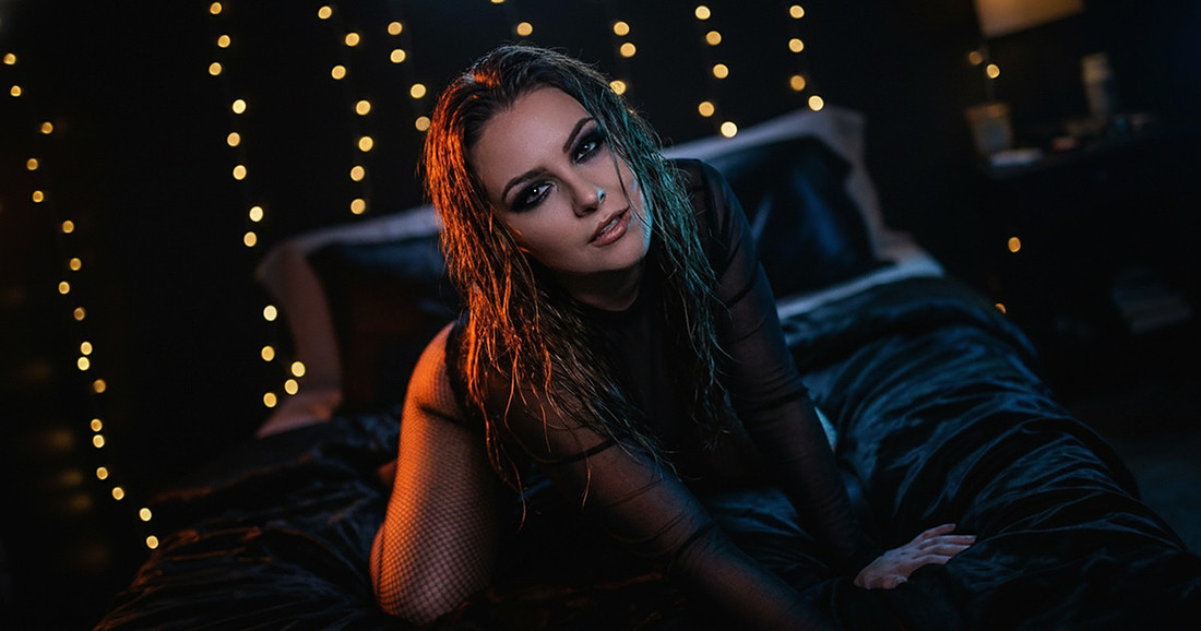 Woman in black lingerie posing on a bed with dark lighting, showcasing intimate boudoir poses and a strong gaze.