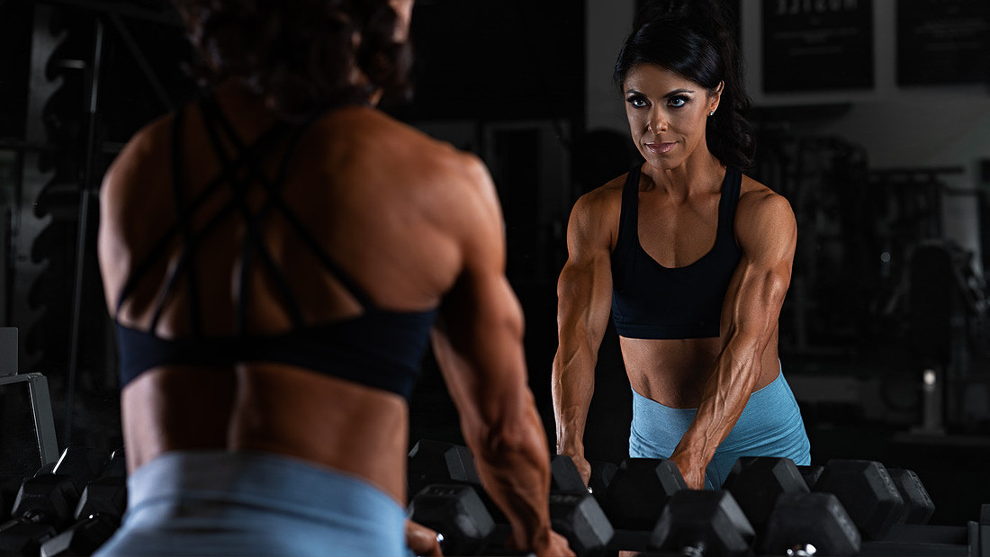 Fitness photoshoot idea of a female bodybuilder racking dumbbells in front of a gym mirror.